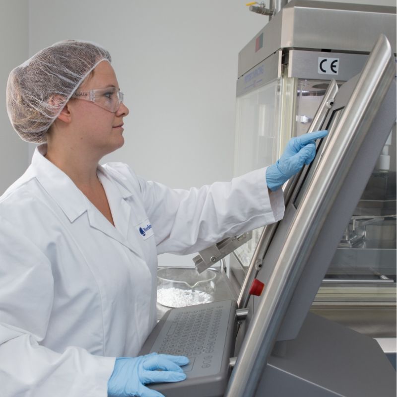 Startseite 38 Eine Frau in Schutzkleidung bedient ein großes Touch-Display an einer Industrieanlage. Sie trägt Laborkittel, Haarnetz, Schutzbrille und blaue Handschuhe.