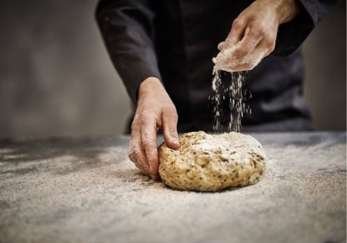 Eine Person bestreut einen Brotteig auf einer bemehlten Arbeitsfläche mit Mehl. Der Teig wird mit beiden Händen bearbeitet.