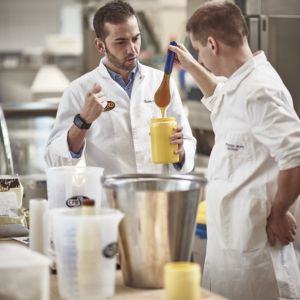 Zwei Männer in weißen Arbeitskitteln stehen in einer Küche. Einer hält ein Glas mit gelber Creme, der andere zieht mit einem Spatel eine Portion davon heraus.